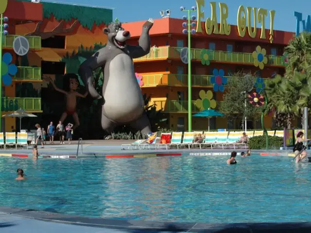 In this picture we see a swimming pool with blue water and people in it. Far away we can see the...